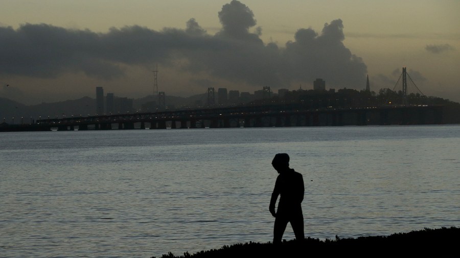 Image: A woman walks along a path as clouds hang over the San Francisco-Oakland Bay Bridge in Emery...